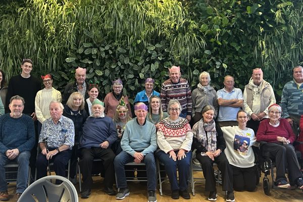 A large group of adults posed for a photo indoors in front of a tall wall covered with dense green plants. The group includes men and women, and various ages. Several people are seated in the front row on chairs, while others stand behind them. They are smiling and wearing casual winter clothing such as sweaters, scarves, and jackets; a few have festive Christmas-style jumpers. Several people are wearing colorful paper party crowns (like Christmas crackers), including pink, purple, green, and red. Two mobility aids are visible in the front-right area: a wheelchair and a rolling walker.