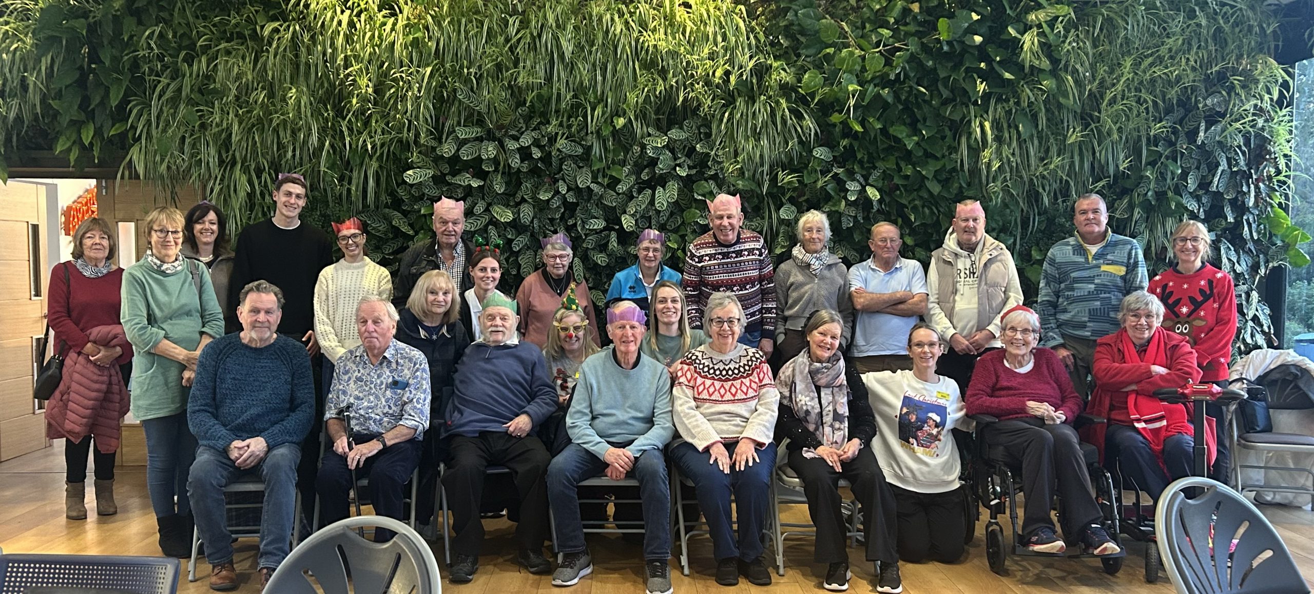A large group of adults posed for a photo indoors in front of a tall wall covered with dense green plants. The group includes men and women, and various ages. Several people are seated in the front row on chairs, while others stand behind them. They are smiling and wearing casual winter clothing such as sweaters, scarves, and jackets; a few have festive Christmas-style jumpers. Several people are wearing colorful paper party crowns (like Christmas crackers), including pink, purple, green, and red. Two mobility aids are visible in the front-right area: a wheelchair and a rolling walker.