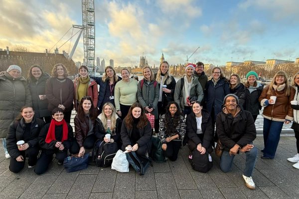 Group of 23 smiling people outside in winter. The London Eye, Thames and Palace of Westminster are in the background.