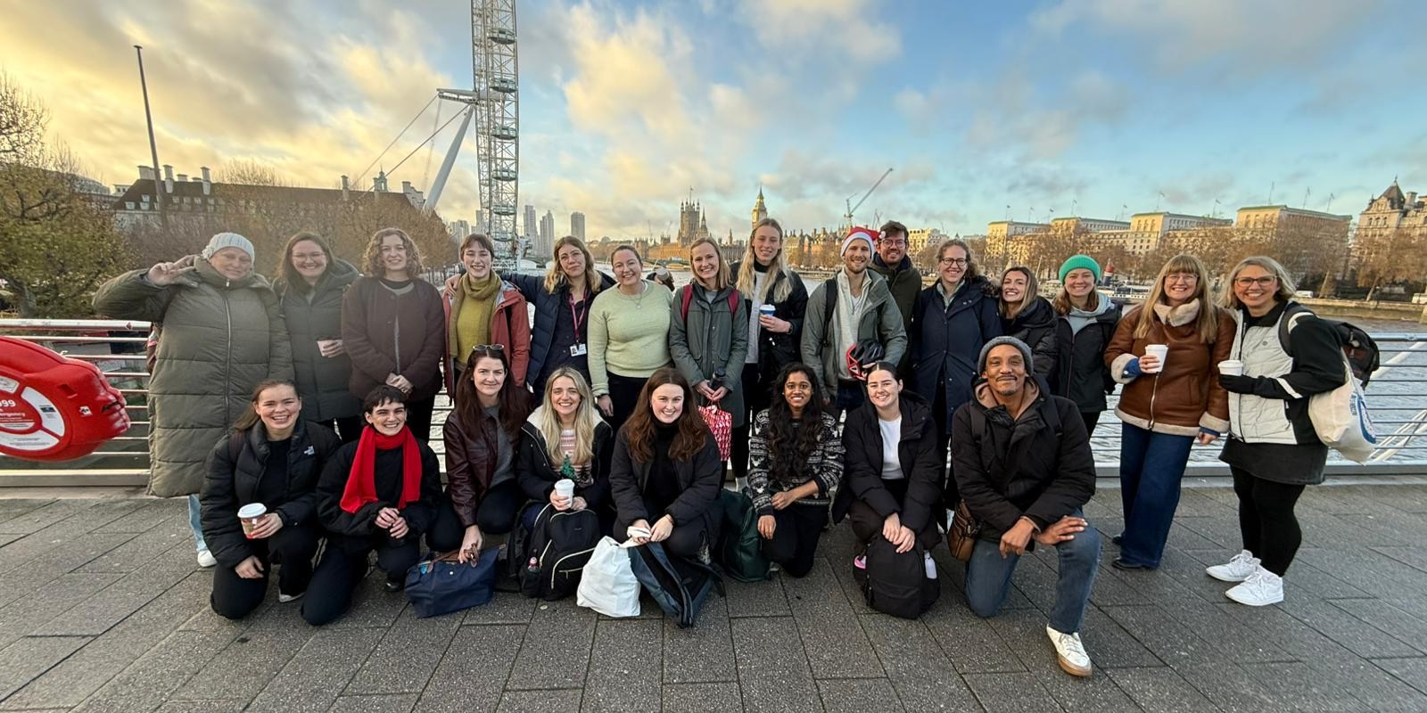 Group of 23 smiling people outside in winter. The London Eye, Thames and Palace of Westminster are in the background.